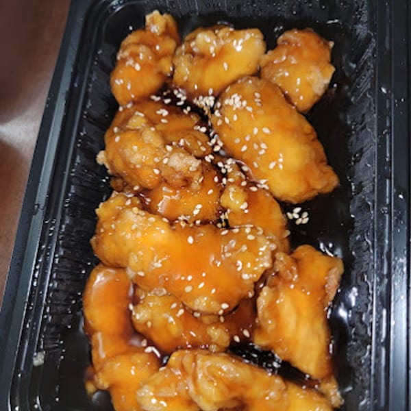 Bao Bao Chinese Bistro's sesame chicken, glistening with sauce and sprinkled with sesame seeds in a black takeout container.