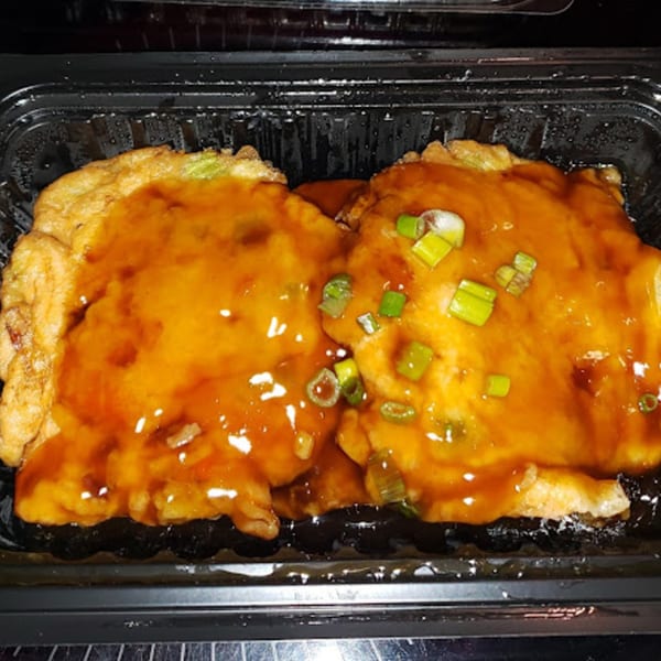 Two golden-brown egg foo young patties covered in a rich brown sauce and green onions in a black container.